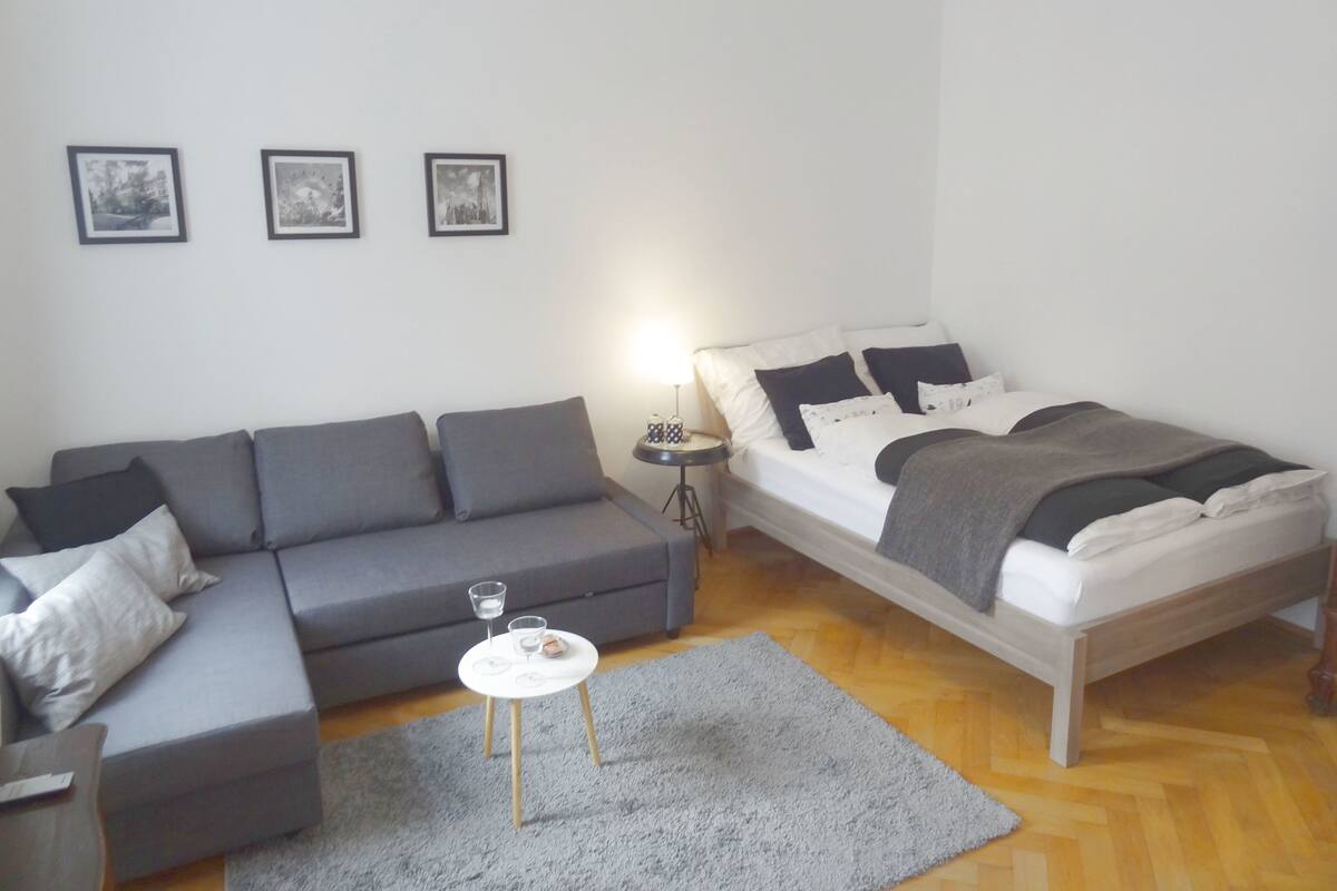A modern bedroom with a grey sofa, a small round table with two glasses, and a bed with a grey blanket.