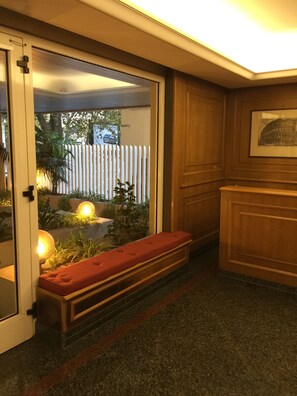 Una habitación con un banco, una puerta que da al exterior, un cuadro enmarcado en la pared y un pequeño jardín visible a través de la puerta.