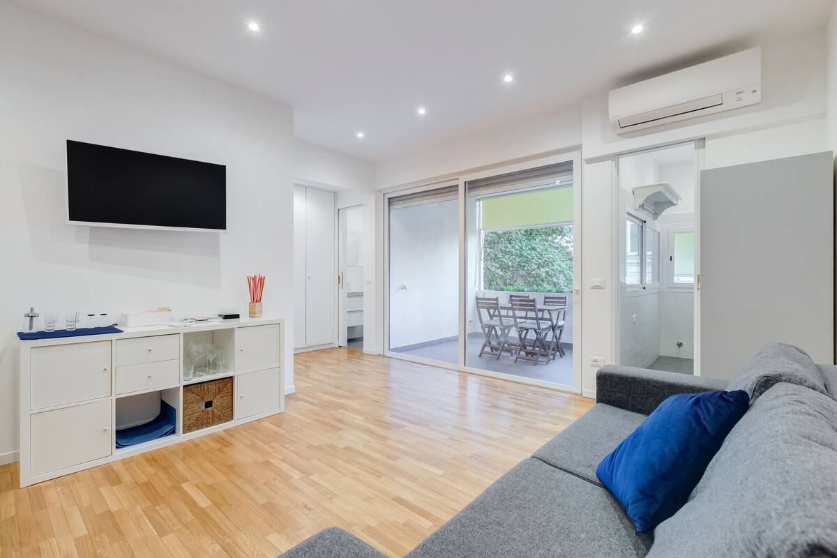 A modern living room with a grey sofa, a flat-screen TV mounted on the wall, a wooden floor, and a sliding glass door leading to an outdoor area with a table and chairs.