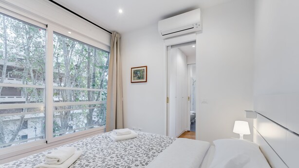 A modern hotel room with a large window, a bed with a patterned bedspread, a bedside table with a lamp, and an air conditioning unit. - Giulio Cesare Luxury Apartment (Rome)