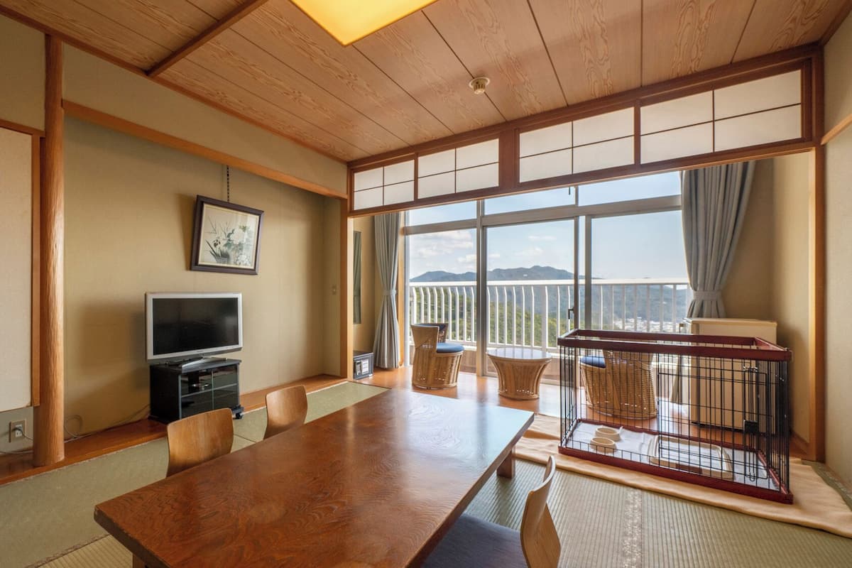 A traditional Japanese-style room with tatami flooring, a wooden table, a television, and a balcony with a view.