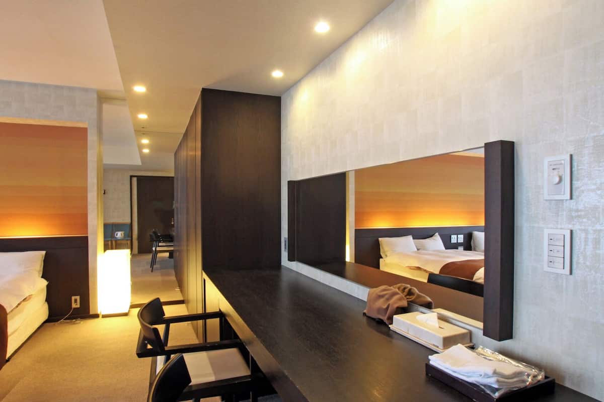 A hotel room with a bed, a desk, a chair, a mirror, and a bedside table with a tissue box and a robe.