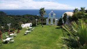 Un césped verde y exuberante con mesas y sillas blancas, un edificio blanco con un campanario y vistas al océano.