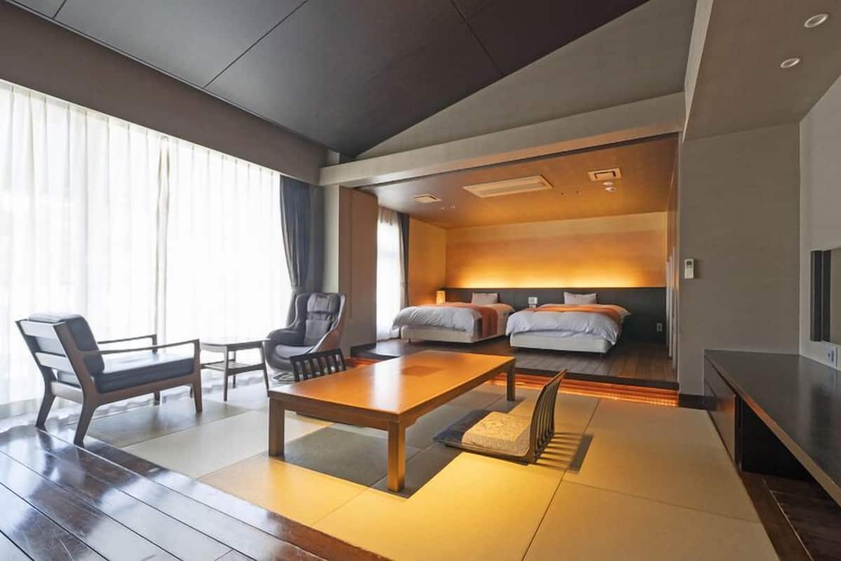 A modern hotel room with a wooden floor, a dining table, two beds, a TV, and a large window with curtains.