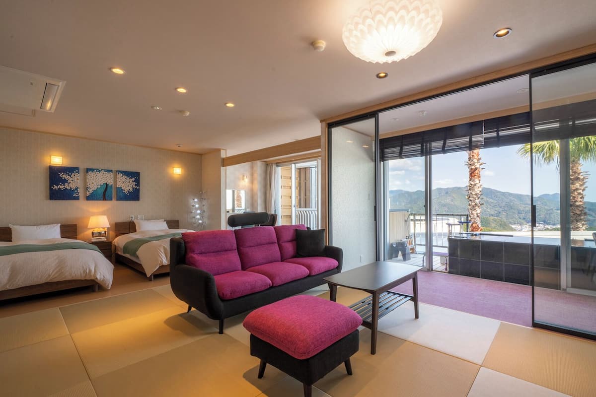A hotel room with a bed, a sofa, a table, and a balcony with a view of the mountains.
