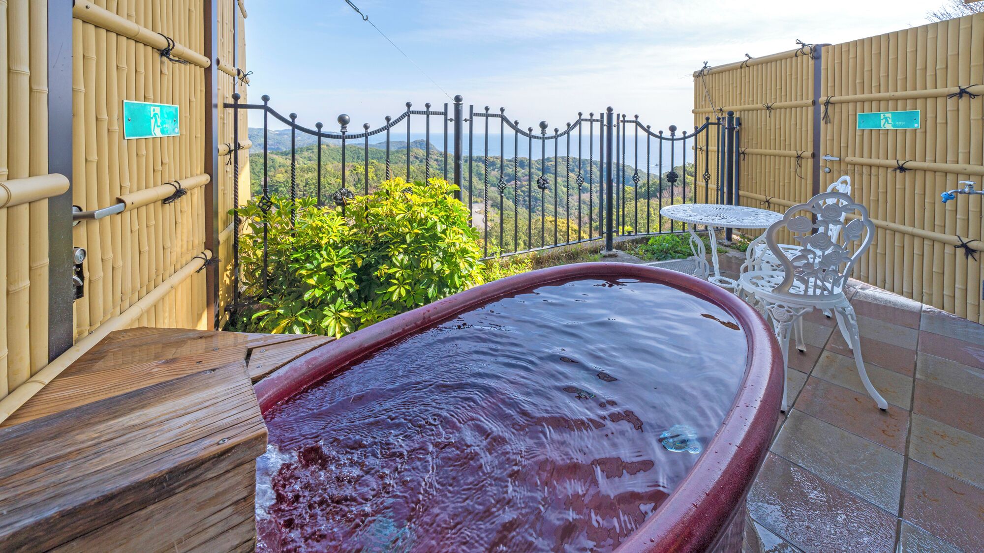 Un bain à remous avec une terrasse en bois, une table blanche avec des chaises et une clôture en bambou offrant une vue sur le paysage.