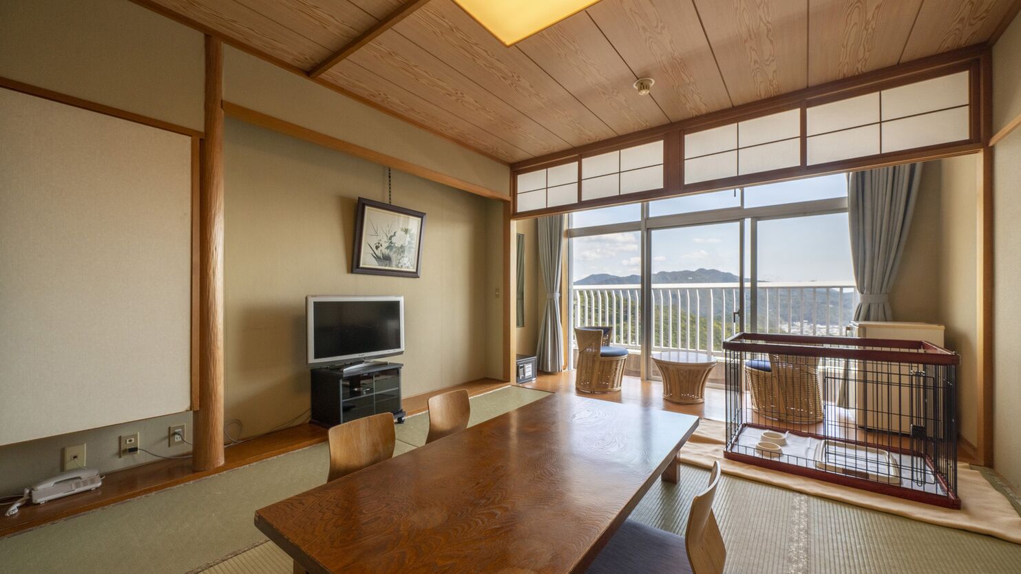 A traditional Japanese-style room with tatami flooring, a wooden table, a television, and a balcony with a view.