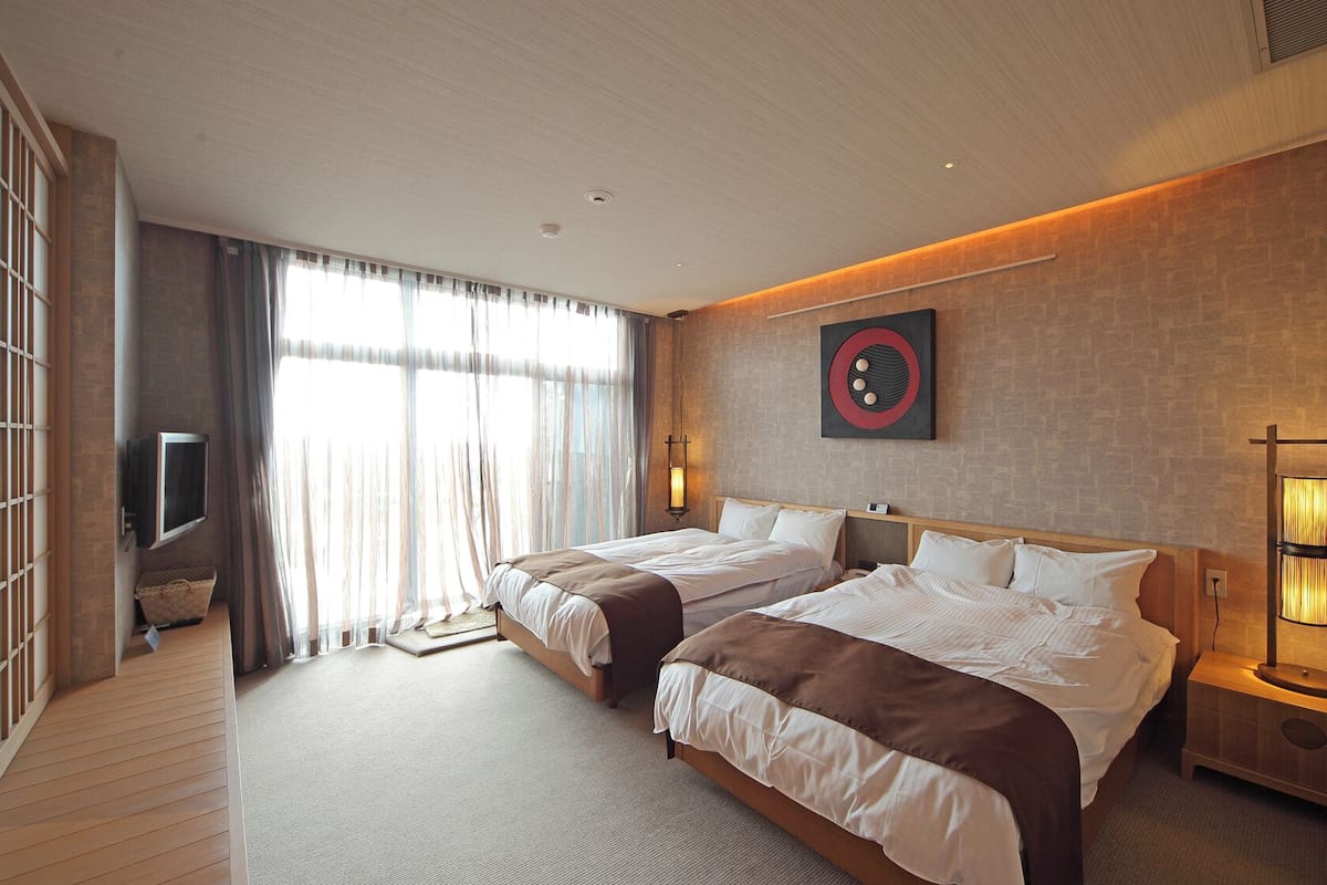 A hotel room with two beds, a television, a wall-mounted artwork, and a bedside table with a lamp.