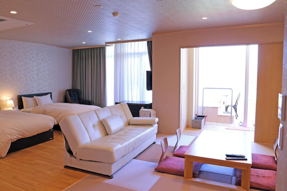 A hotel room with a large bed, a sofa, a dining table, and a view of the outside.