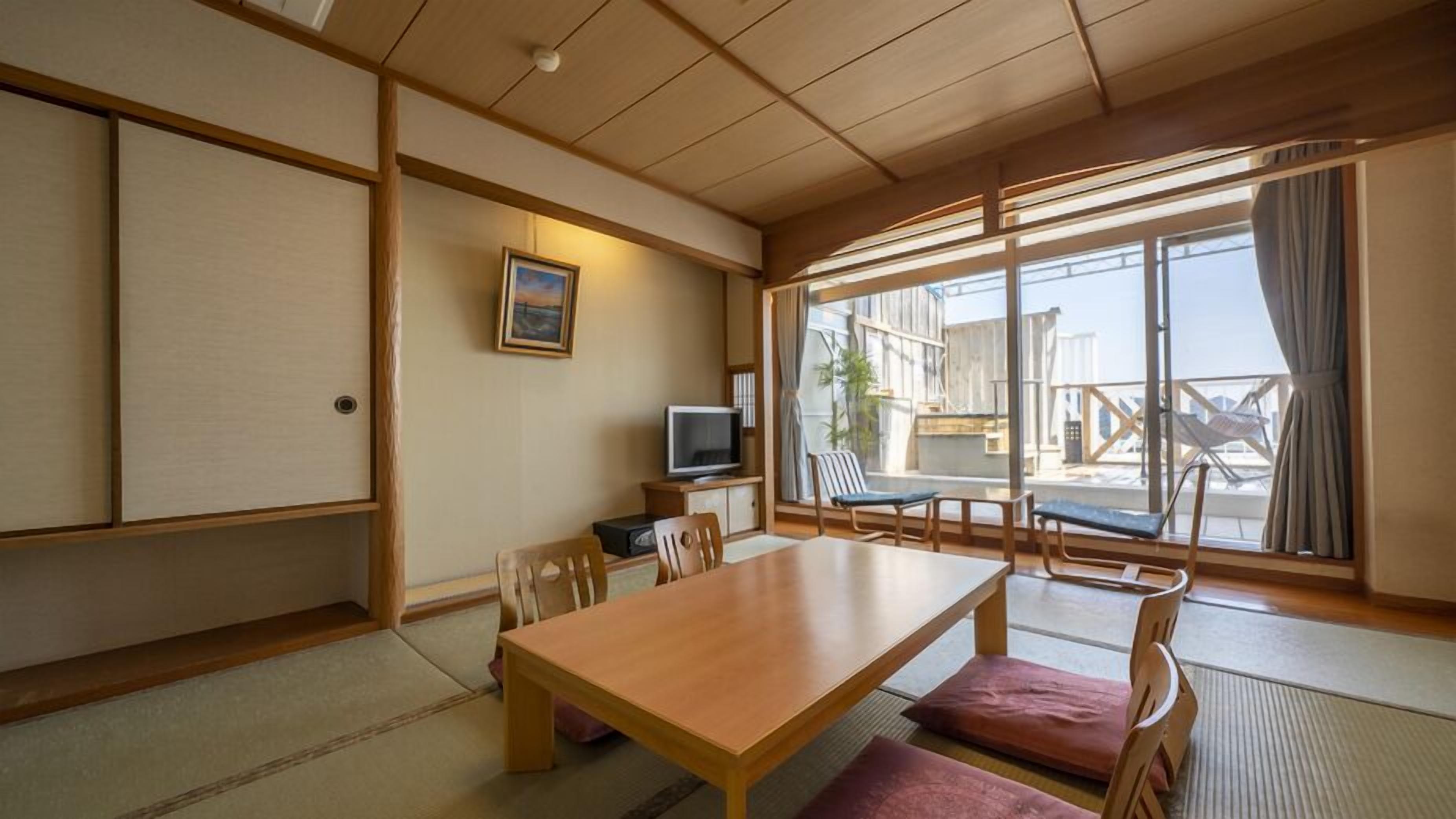 A traditional Japanese room with tatami flooring, a wooden table, chairs, a television, and large windows with a view of the outside.