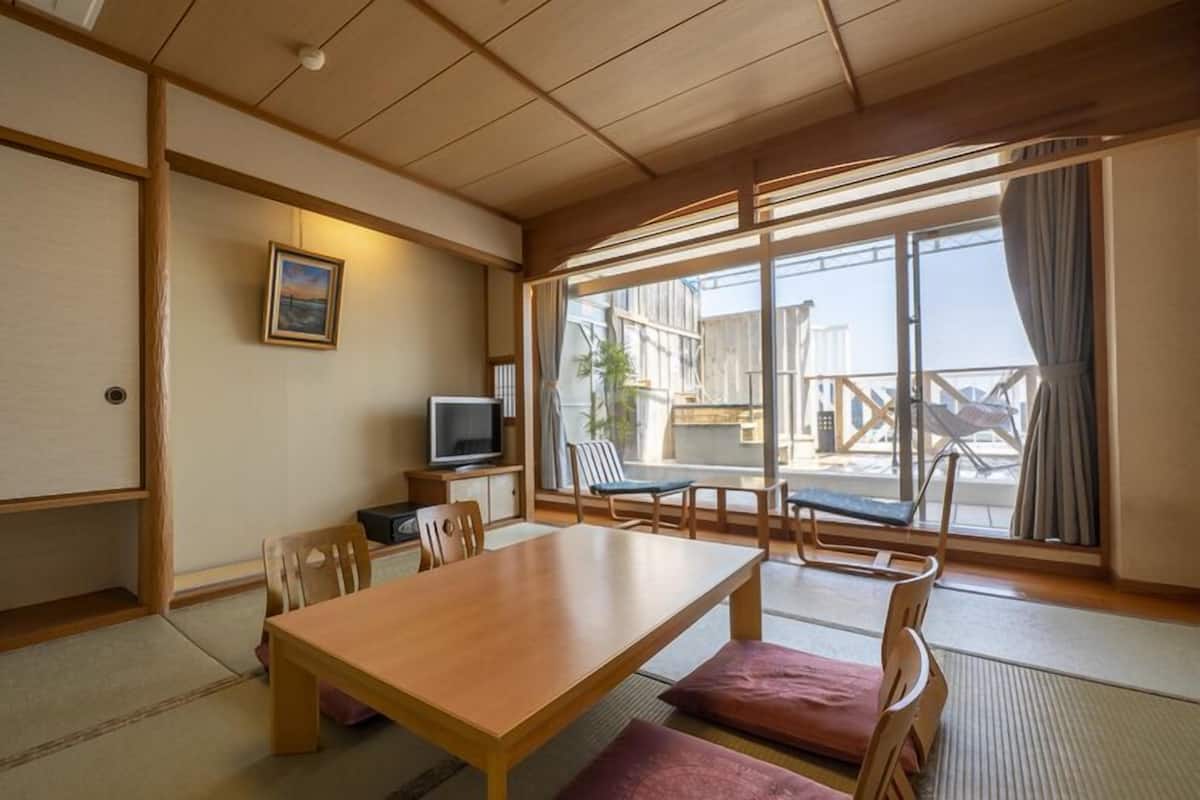 A traditional Japanese room with tatami flooring, a wooden table, chairs, a television, and large windows with a view of the outside.