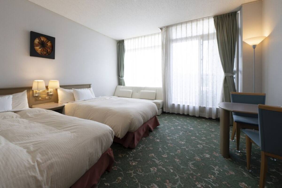 A hotel room with two beds, a desk, a chair, a lamp, and a window with curtains.