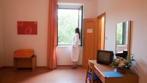 Une pièce avec une fenêtre, un bureau avec un ordinateur, une chaise, une valise et une porte.