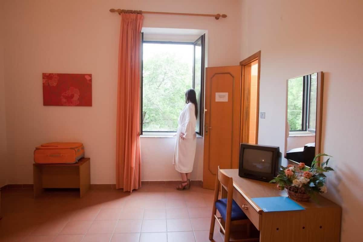 A room with a window, a desk with a computer, a chair, a suitcase, and a door.