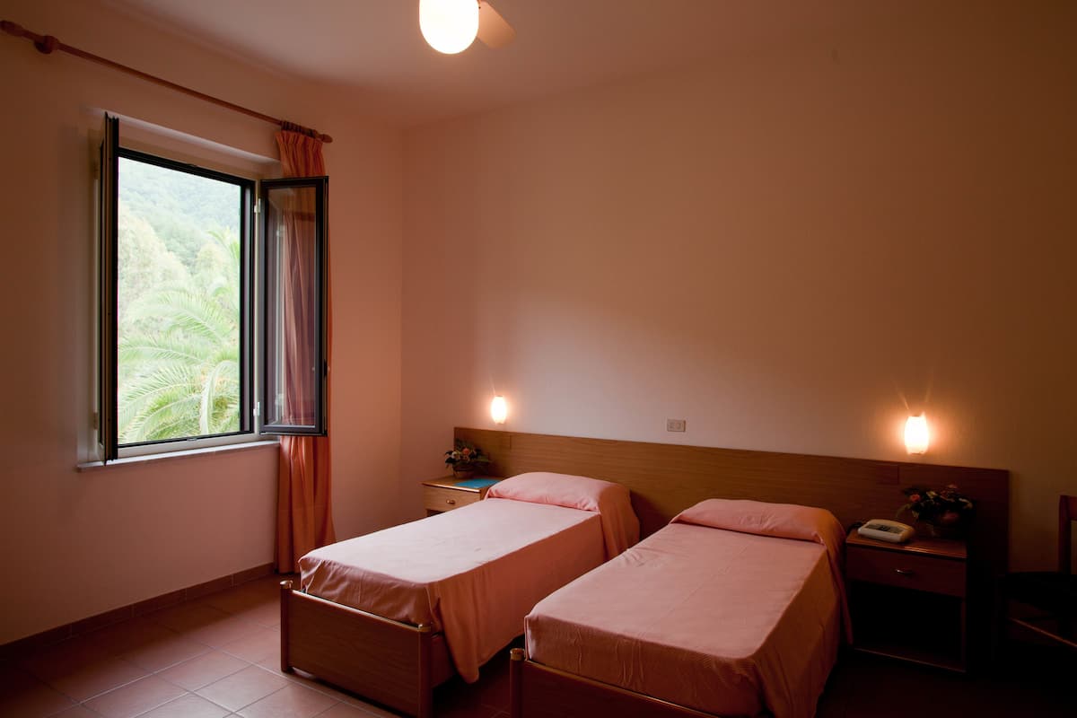 A room with two beds, a window with curtains, a nightstand with a lamp, and a wall-mounted light.