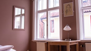 A room with a wooden desk, a chair, a bed with a white bedspread, and a window with a view of a red-roofed building outside. - Adelsgatan Bed & Breakfast (Visby)