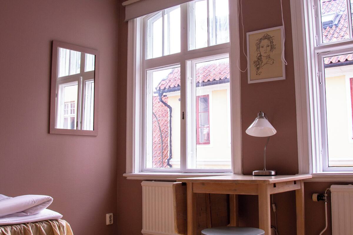 A room with a wooden desk, a chair, a bed with a white bedspread, and a window with a view of a red-roofed building outside.