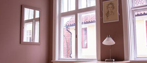 A room with a wooden desk, a chair, a bed with a white bedspread, and a window with a view of a red-roofed building outside.
