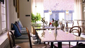 A dining area with a long table, chairs, and a sofa with cushions. There are hanging lights and a window with a view of a building with a tiled roof. - Adelsgatan Bed & Breakfast (Visby)