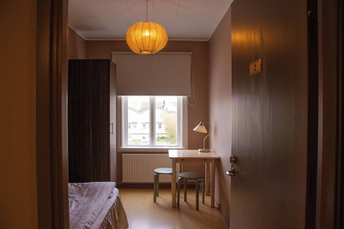 A room with a bed, a desk with a chair, a lamp, and a window with blinds.