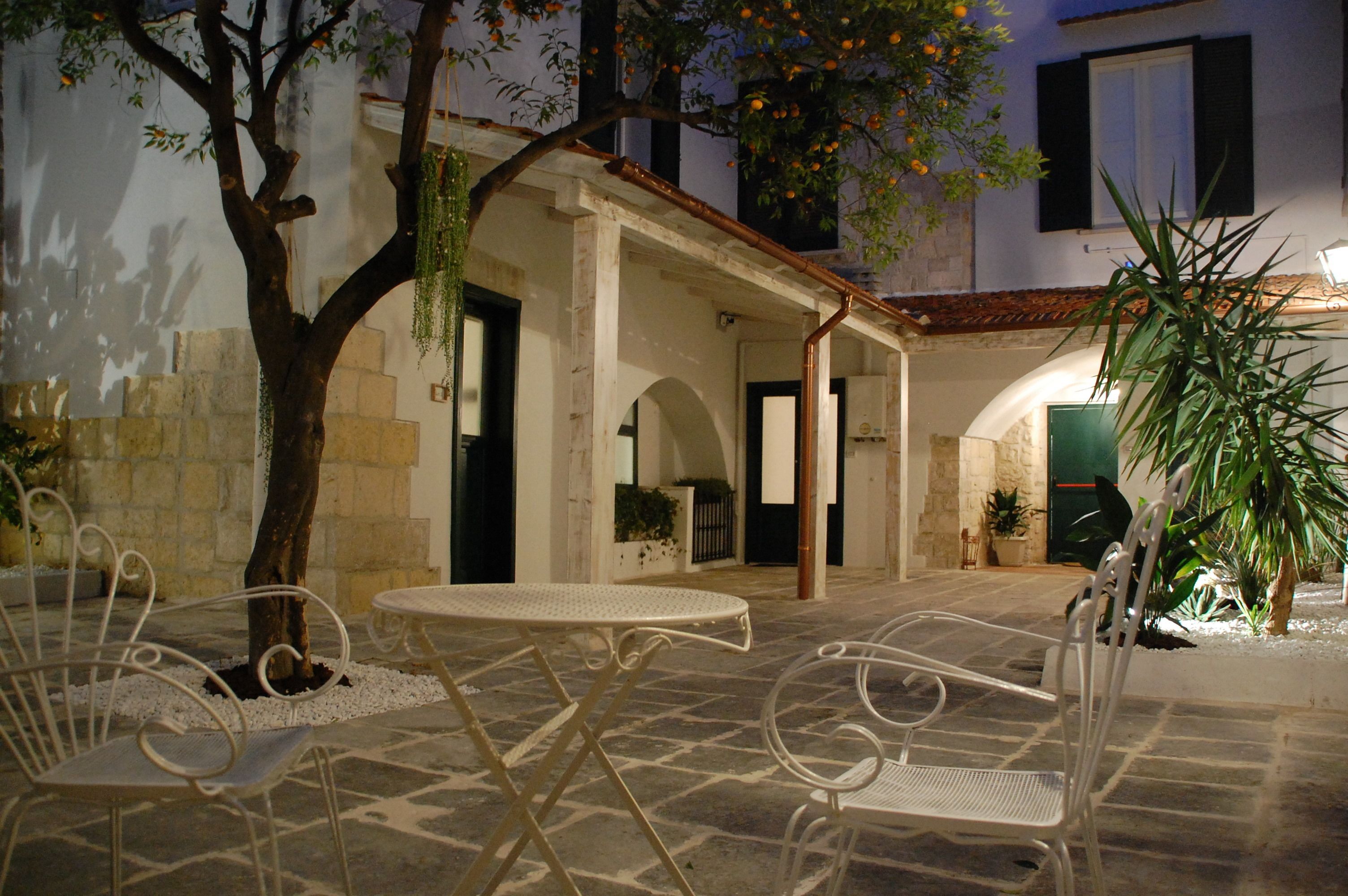 An outdoor patio with stone flooring, white metal chairs, and tables, surrounded by plants and a tree.