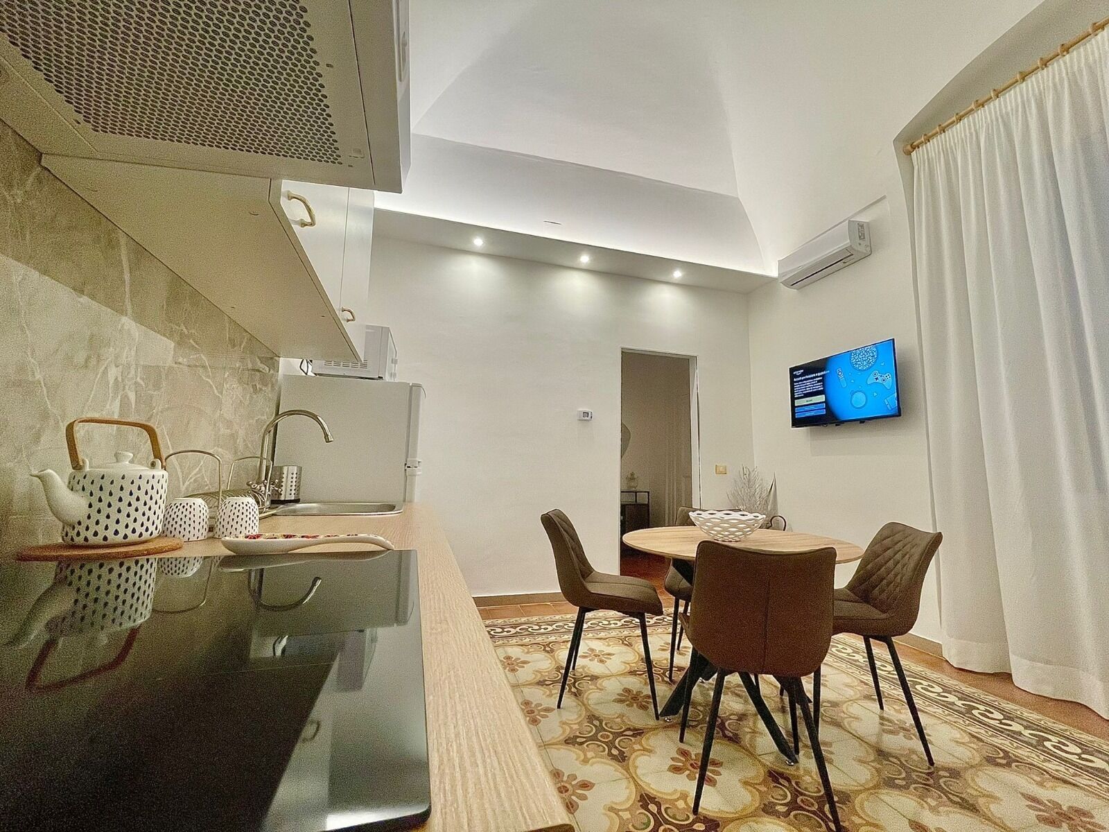 A modern kitchen with a dining area, a TV mounted on the wall, and a patterned rug.