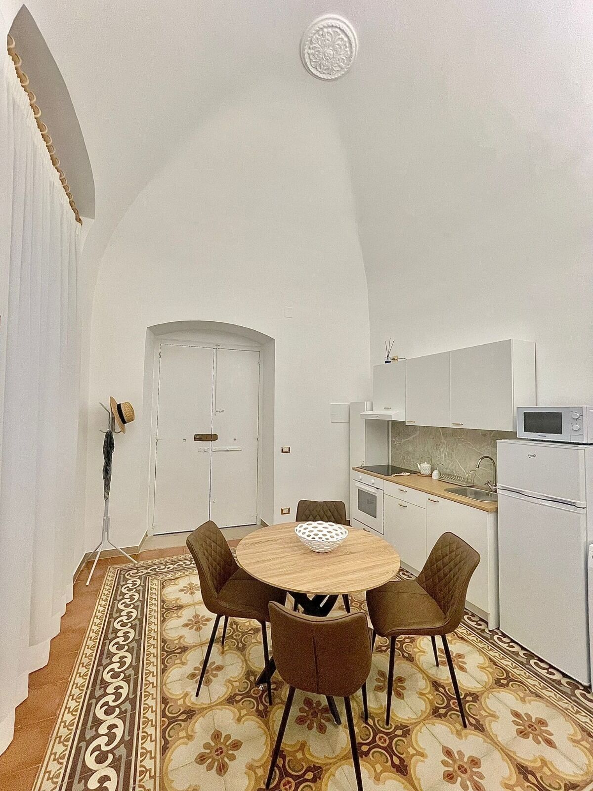 A compact kitchen with white cabinets, a microwave, and a refrigerator. A dining table with four chairs is in the center, surrounded by a patterned floor.