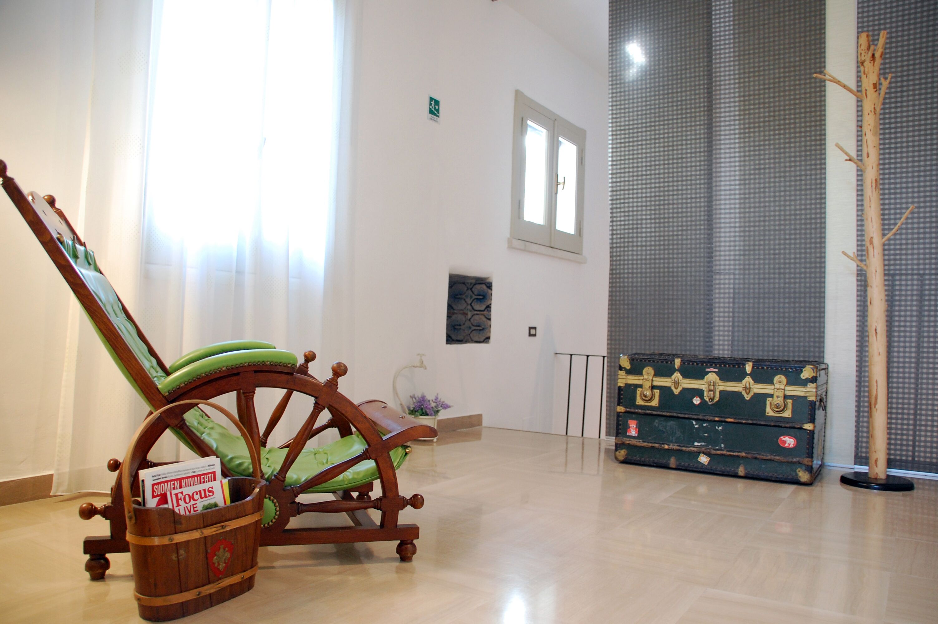 Una stanza con una sedia a dondolo in legno, una poltrona imbottita verde, un secchio di legno con un cartello e una valigia vintage.