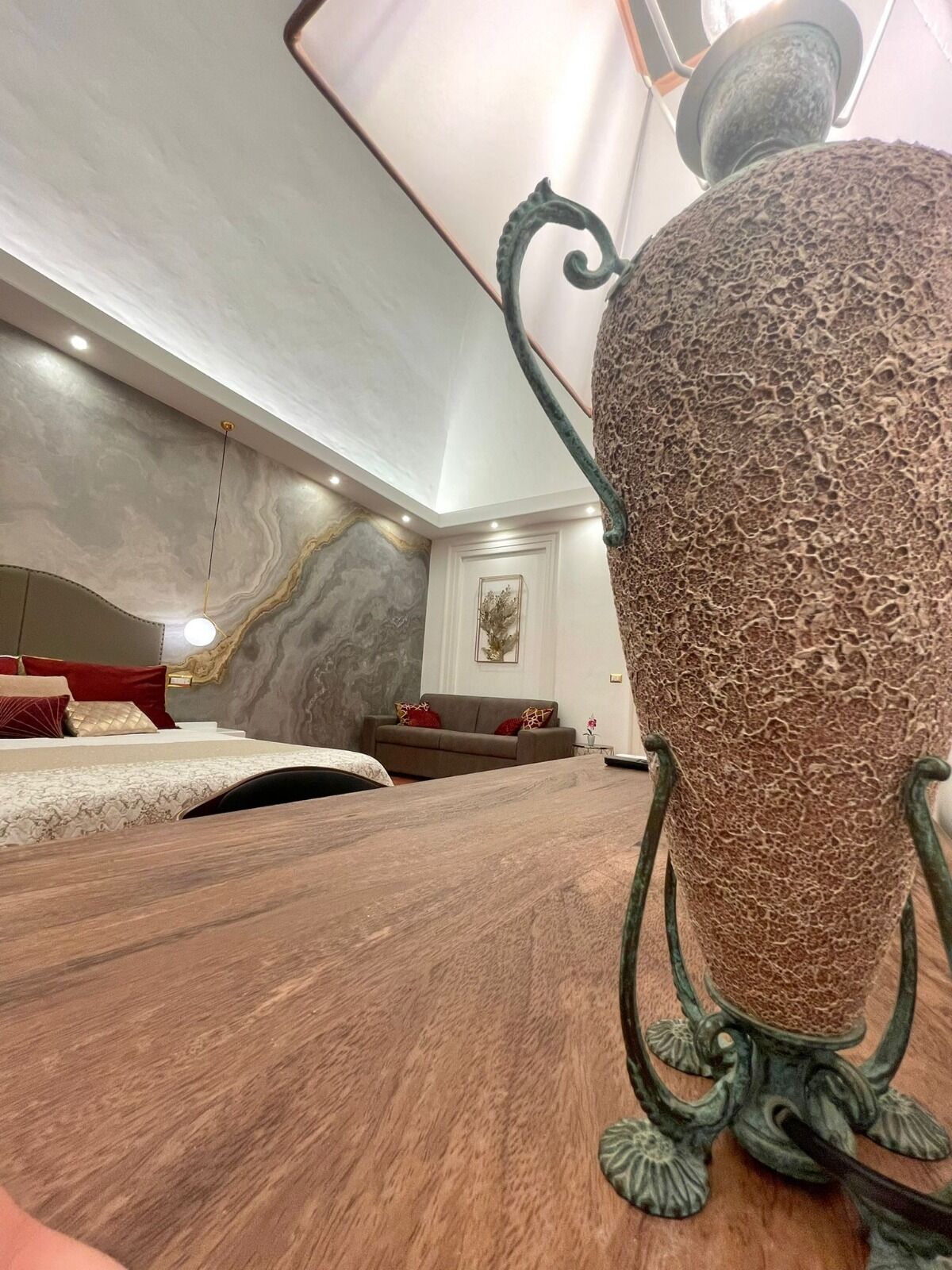 A spacious room with a large, textured vase on a wooden table, a sofa, and a bed with red pillows.