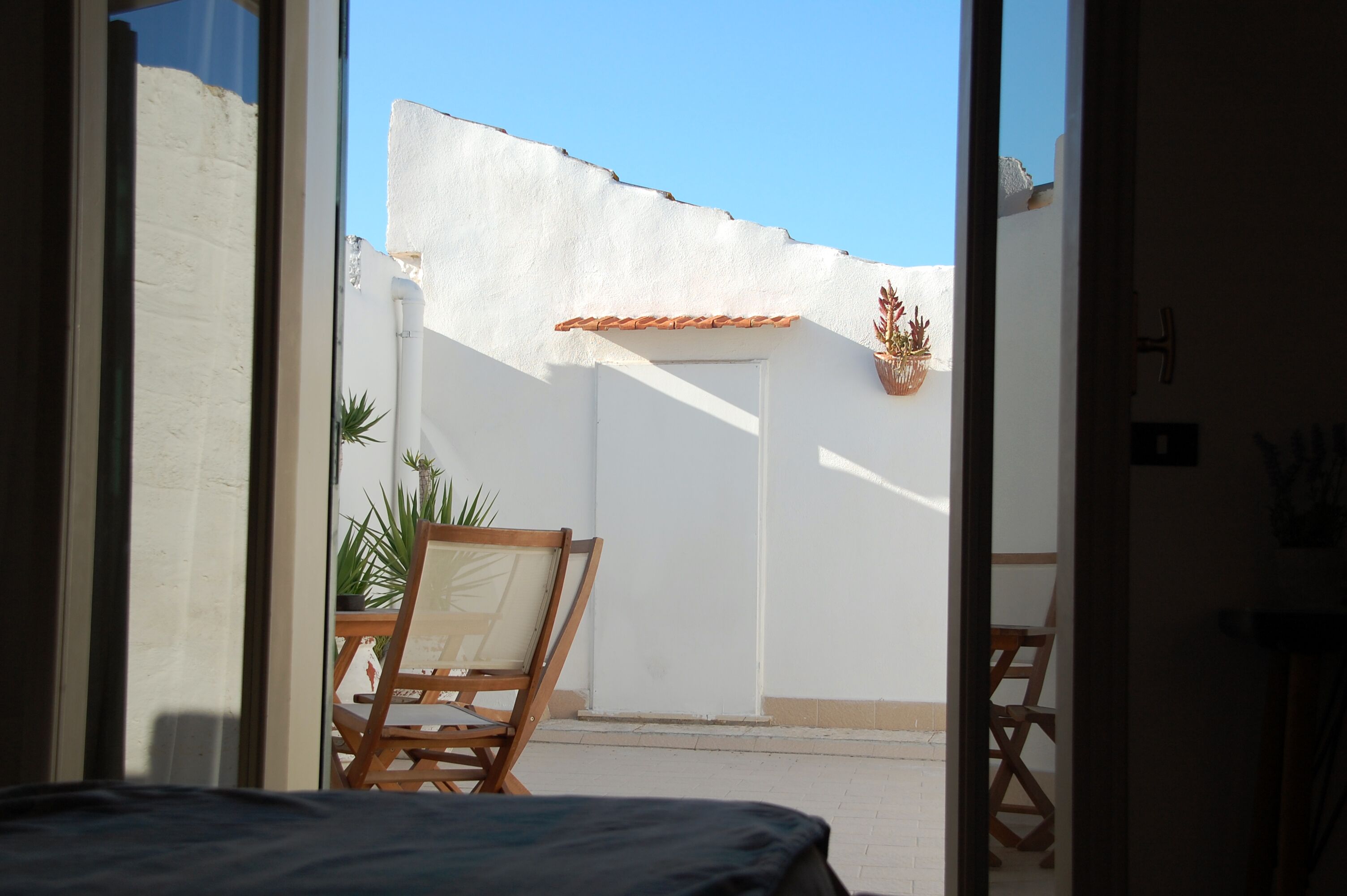 A room with a bed, a wooden chair, a potted plant, and a view of a white wall with a tiled roof.