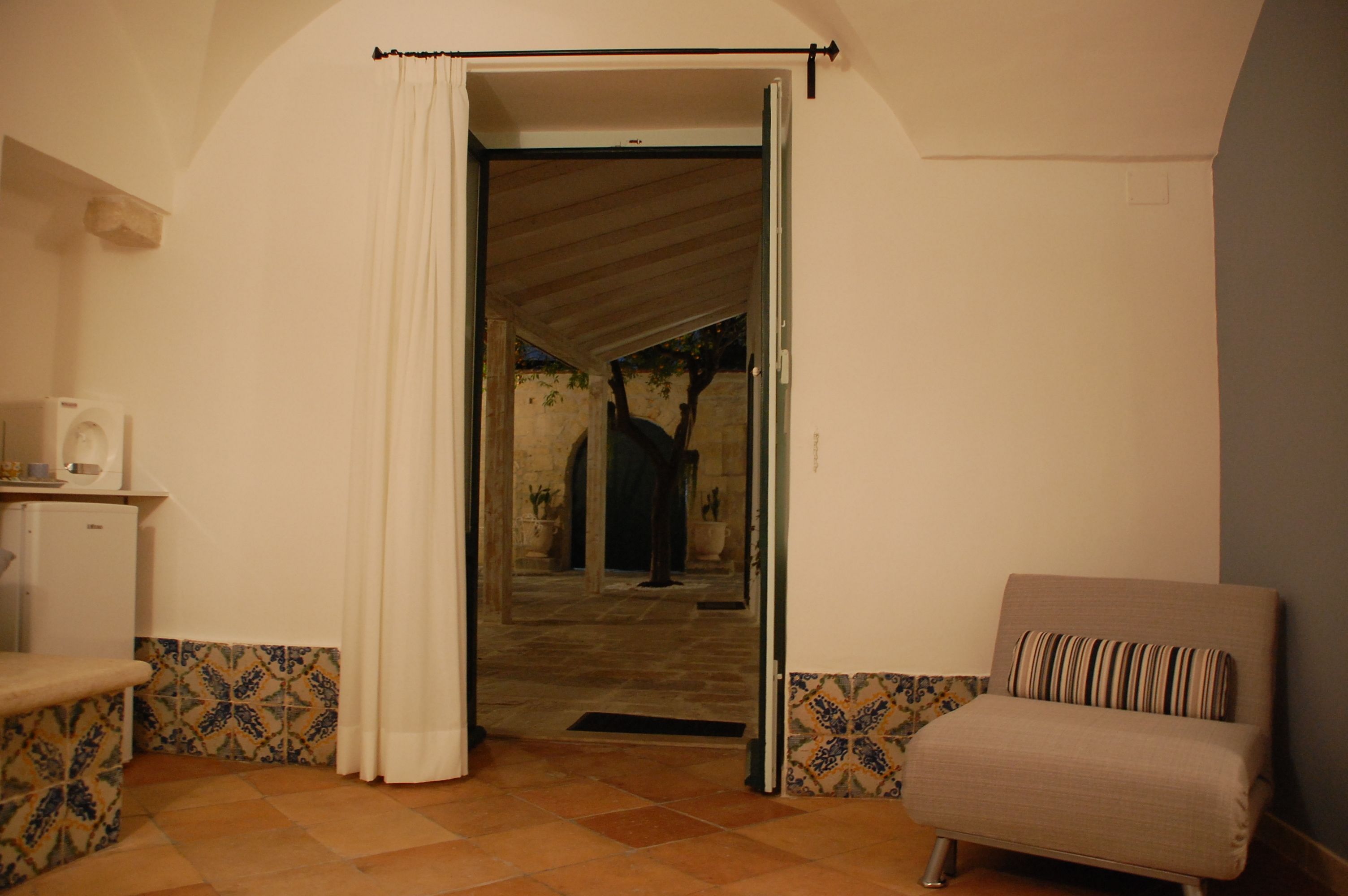 A room with a white door, a small kitchenette, a beige armchair, and patterned floor tiles.