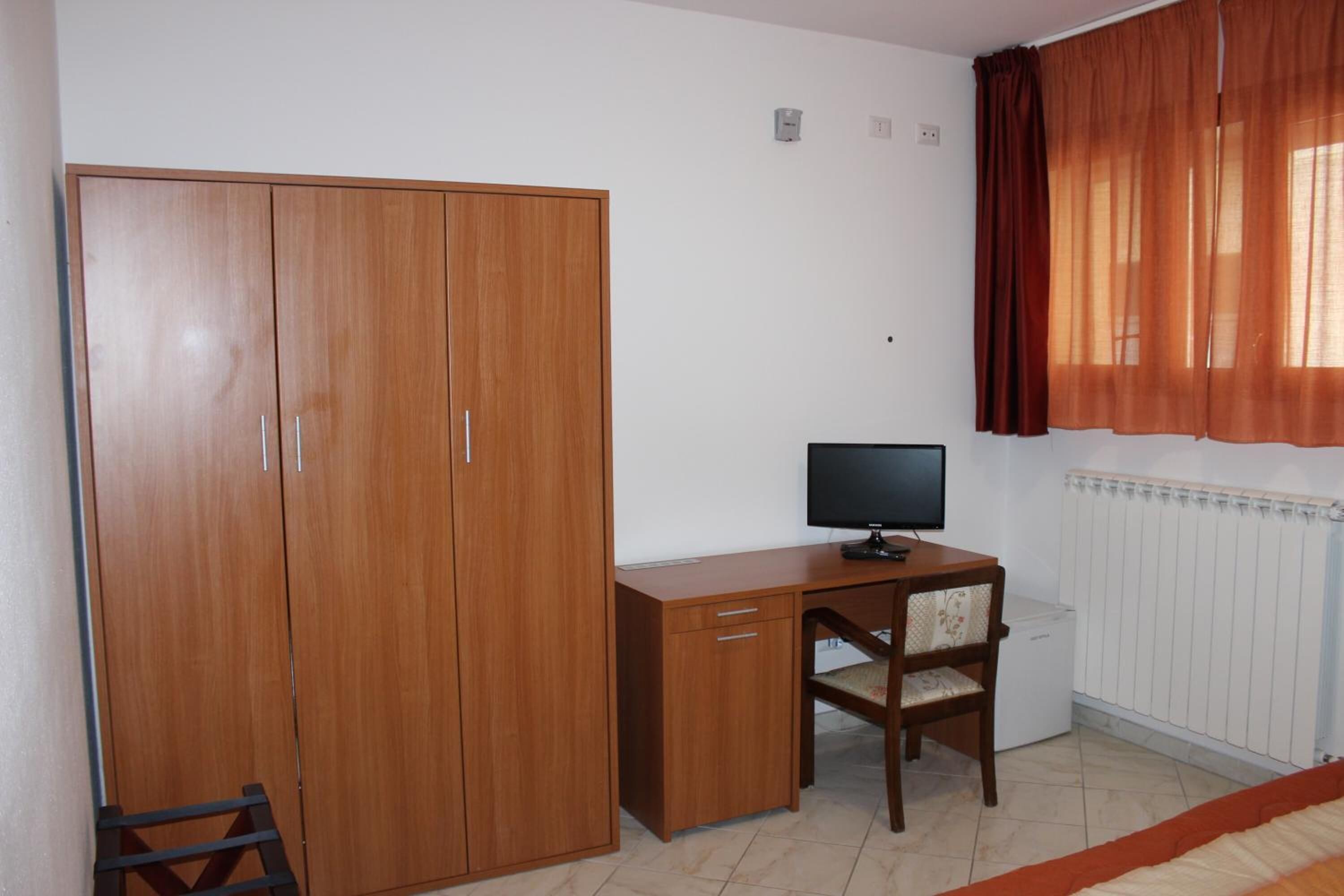 Quarto com guarda-roupa de madeira, escrivaninha com cadeira, televisão e radiador.