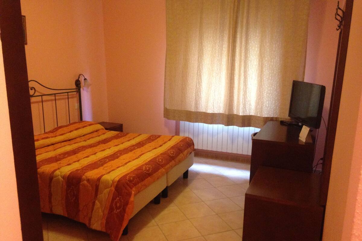 Een slaapkamer met een bed, een televisie op een standaard en een raam met gordijnen.