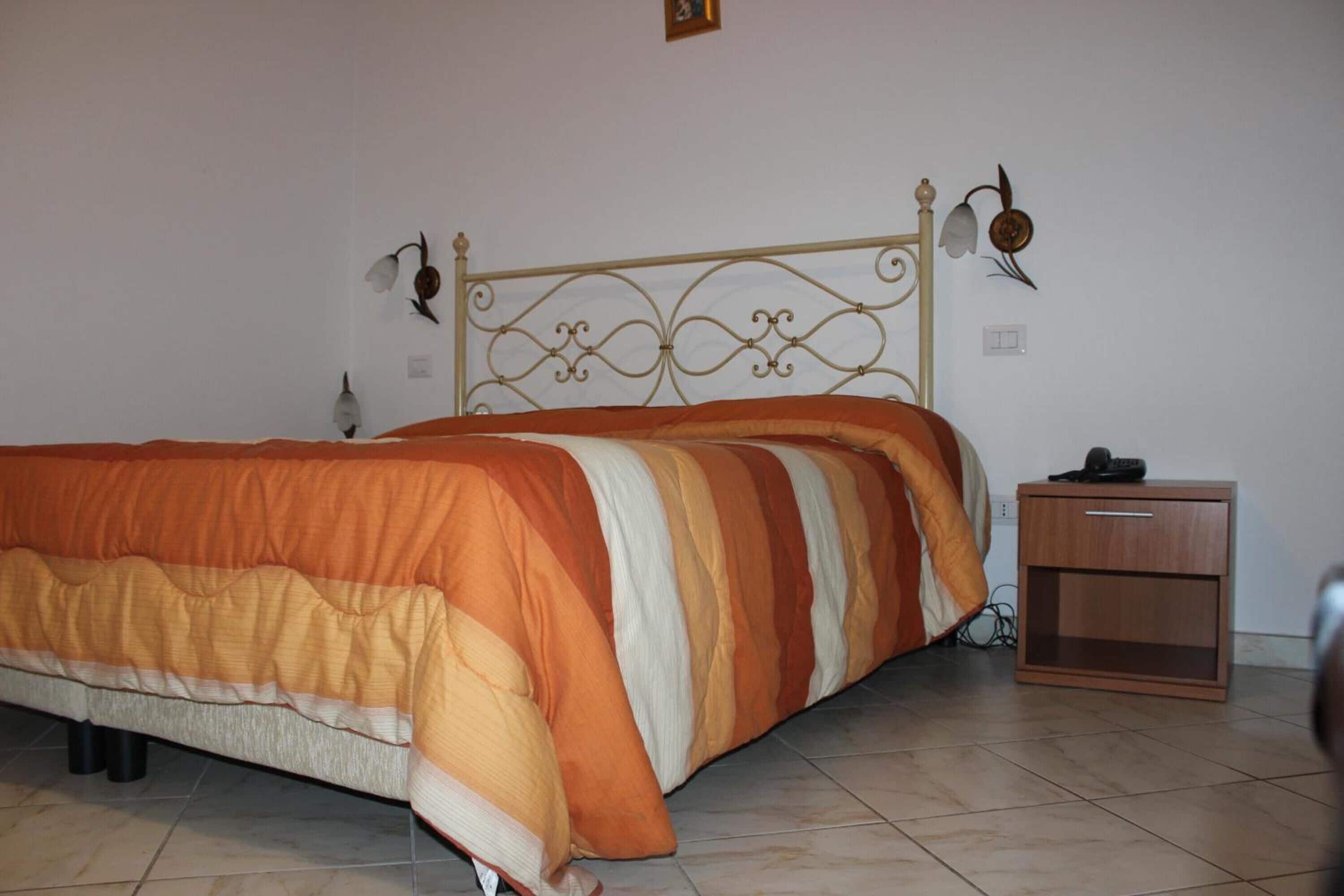 Un lit avec une couverture à rayures, une tête de lit en métal, une table de chevet avec un tiroir et un luminaire fixé au mur.