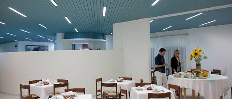 A dining area with tables set for service, wooden chairs, and a ceiling with linear lights.