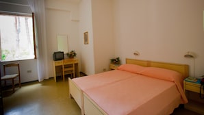 Een slaapkamer met een bed, een bureau met een televisie, een stoel, een nachtkastje met een lamp en een raam met gordijnen.