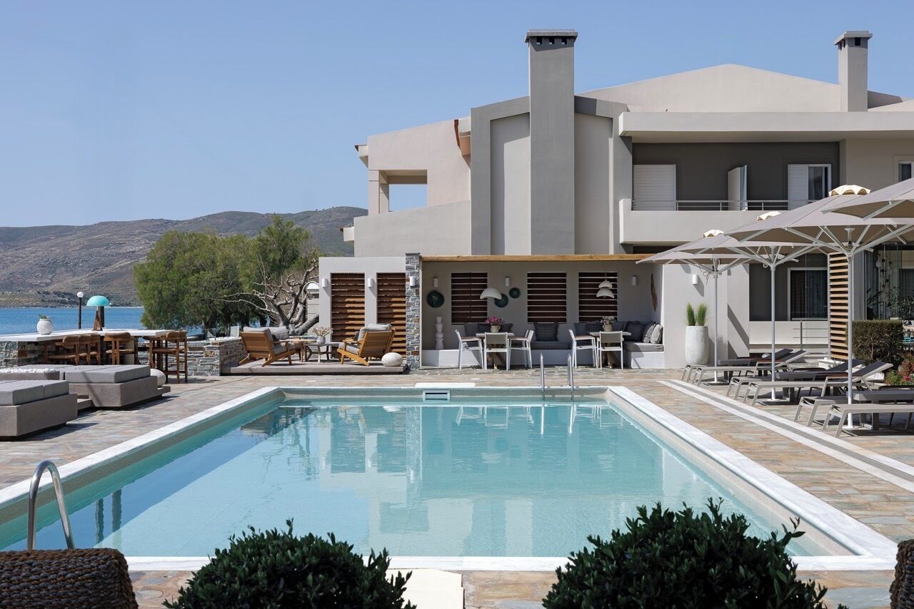 A modern pool area with lounge chairs and umbrellas, a clear view of the sea, and a large building with a flat roof and chimneys.