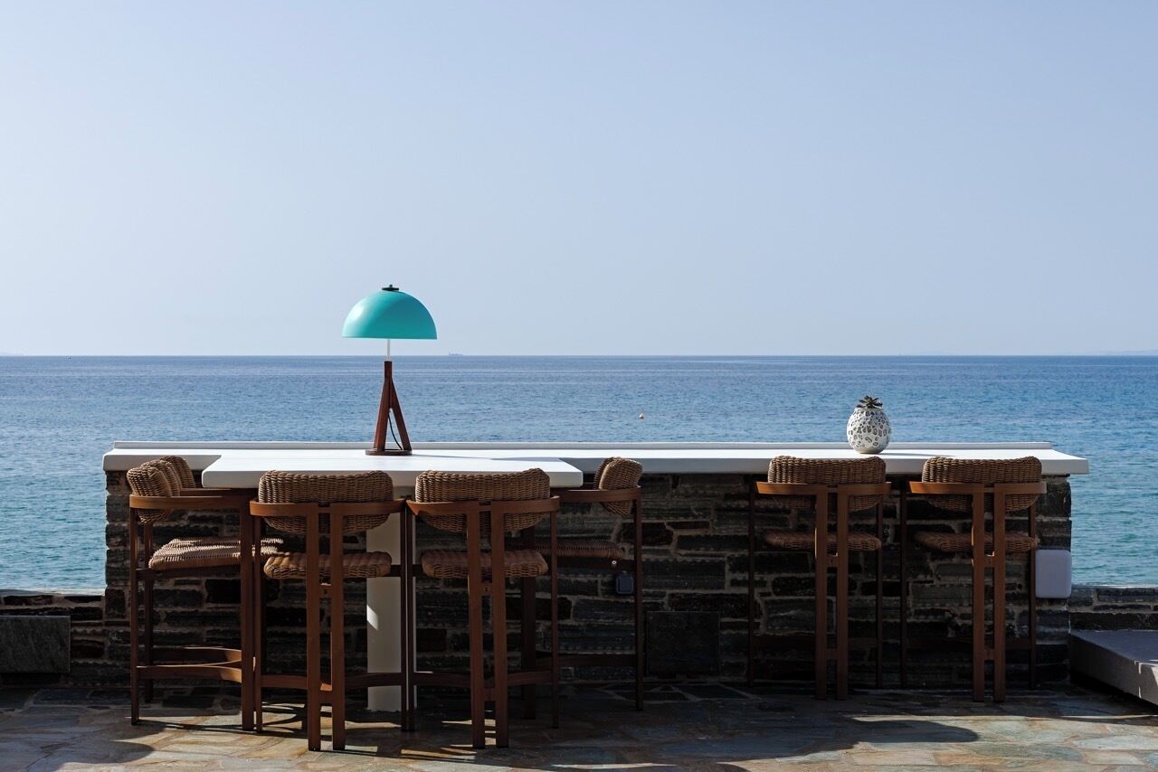 Un bar en bord de mer avec des murs en pierre, un comptoir blanc et des tabourets de bar en osier.