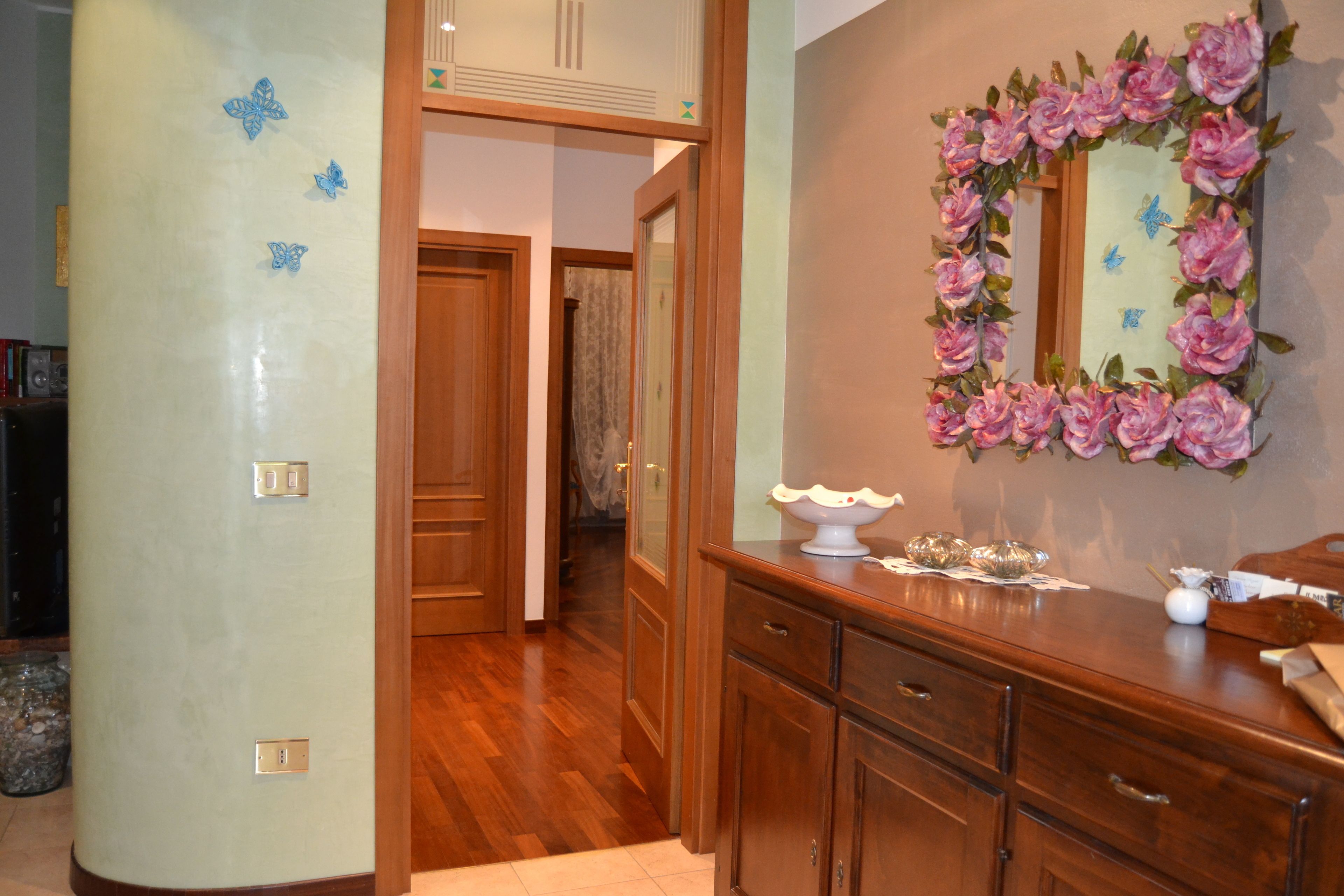 A room with wooden flooring, a wooden dresser, a mirror with a floral frame, and a door leading to another room.