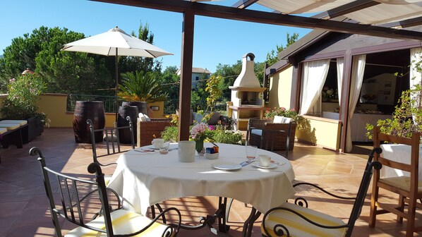 Un patio al aire libre con una mesa redonda dispuesta para comer, rodeado de sillas de mimbre y un paraguas grande.