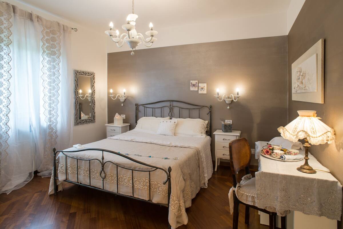 A bedroom with a bed, bedside tables, a chair, a table with a lamp, and a chandelier.