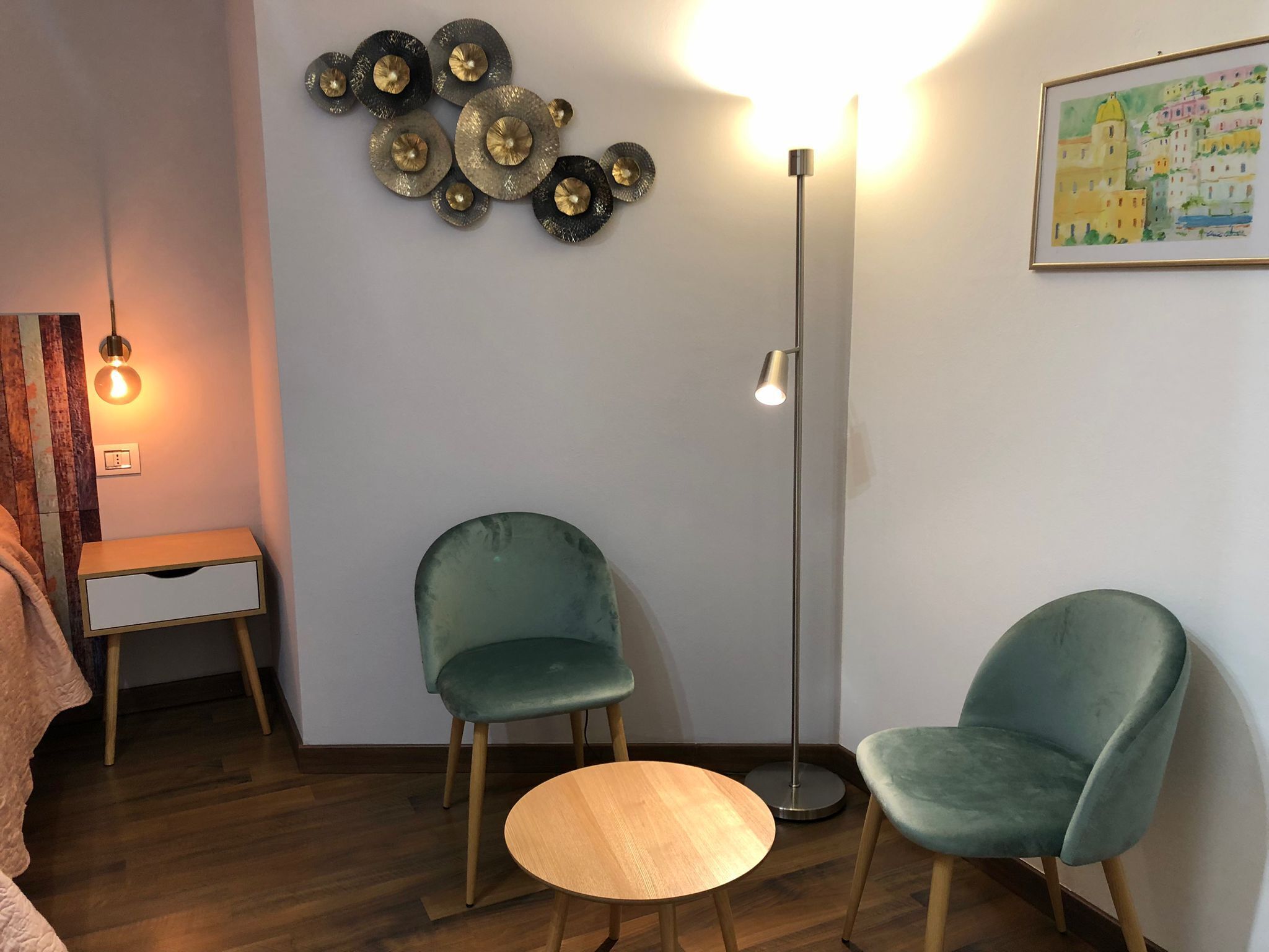 A room with a wooden floor, two green chairs, a small wooden table, a floor lamp, a wall art piece, and a bed with a blanket.