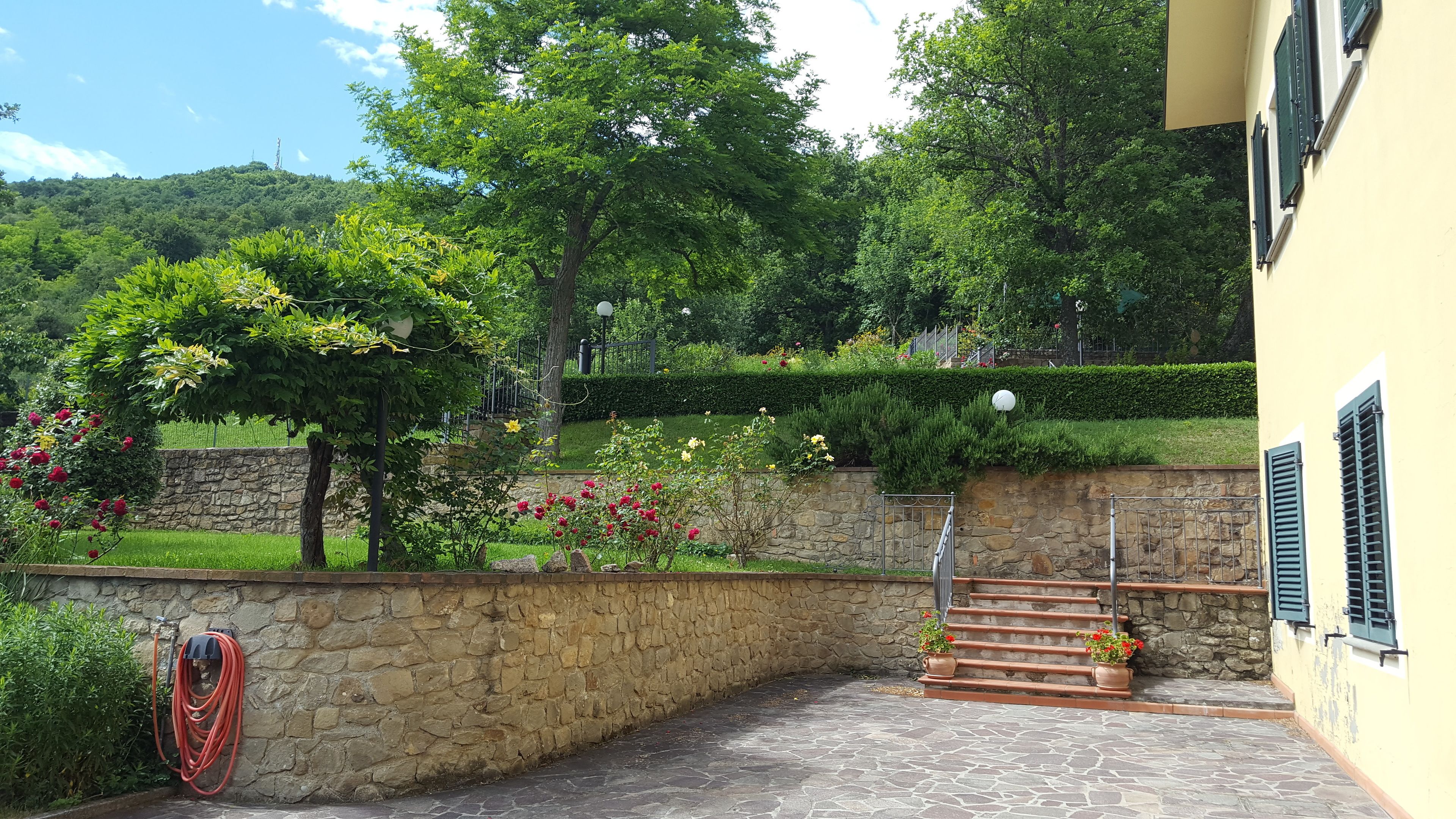 Un mur de pierre bordé d’un jardin, avec un tuyau d’arrosage et un escalier menant à un bâtiment aux volets verts.