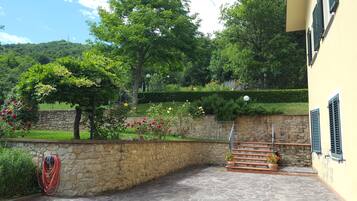 Un mur de pierre bordé d’un jardin, avec un tuyau d’arrosage et un escalier menant à un bâtiment aux volets verts.