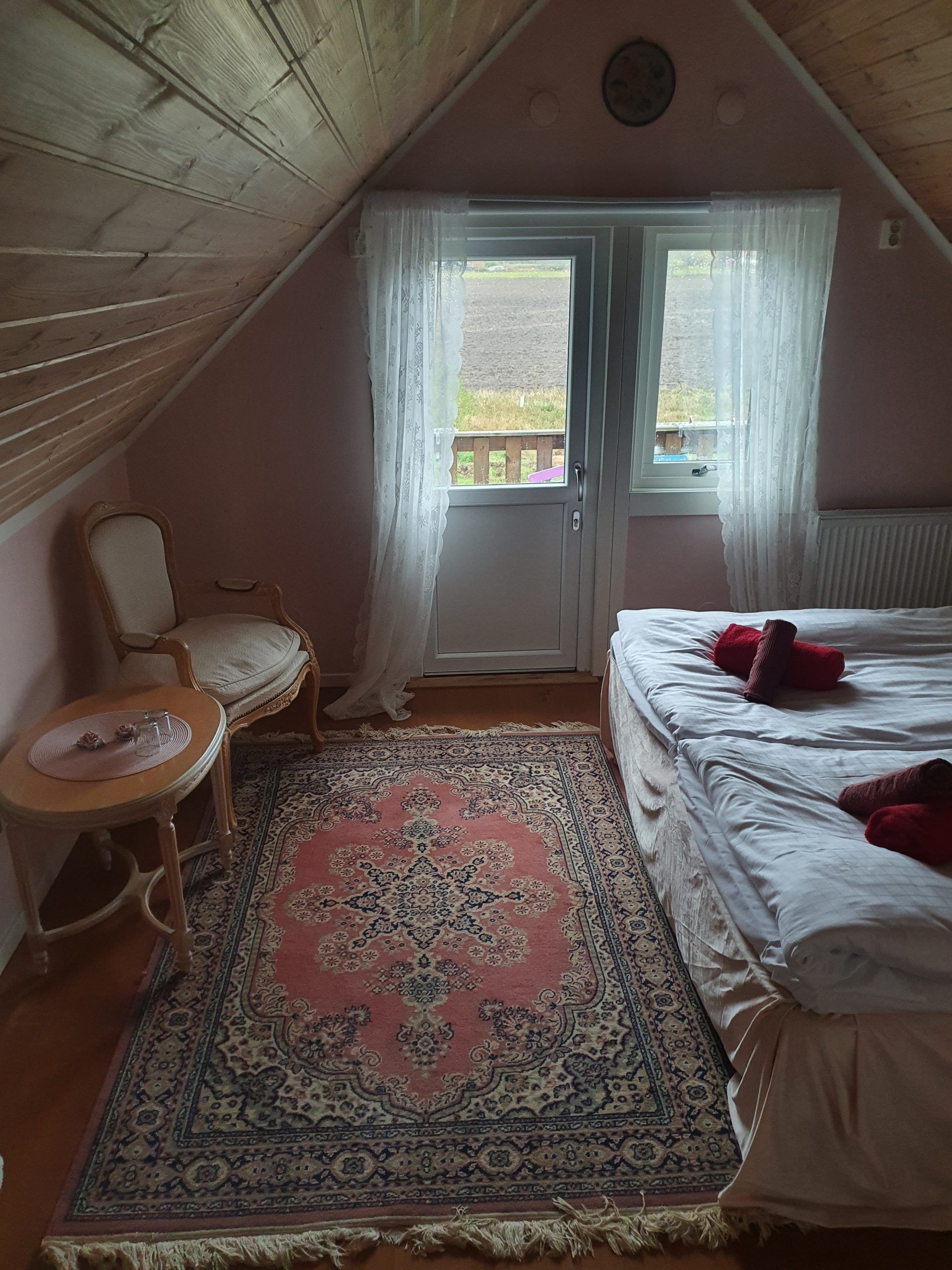 A cozy attic room with a large bed, a chair, a small table, and a patterned rug.