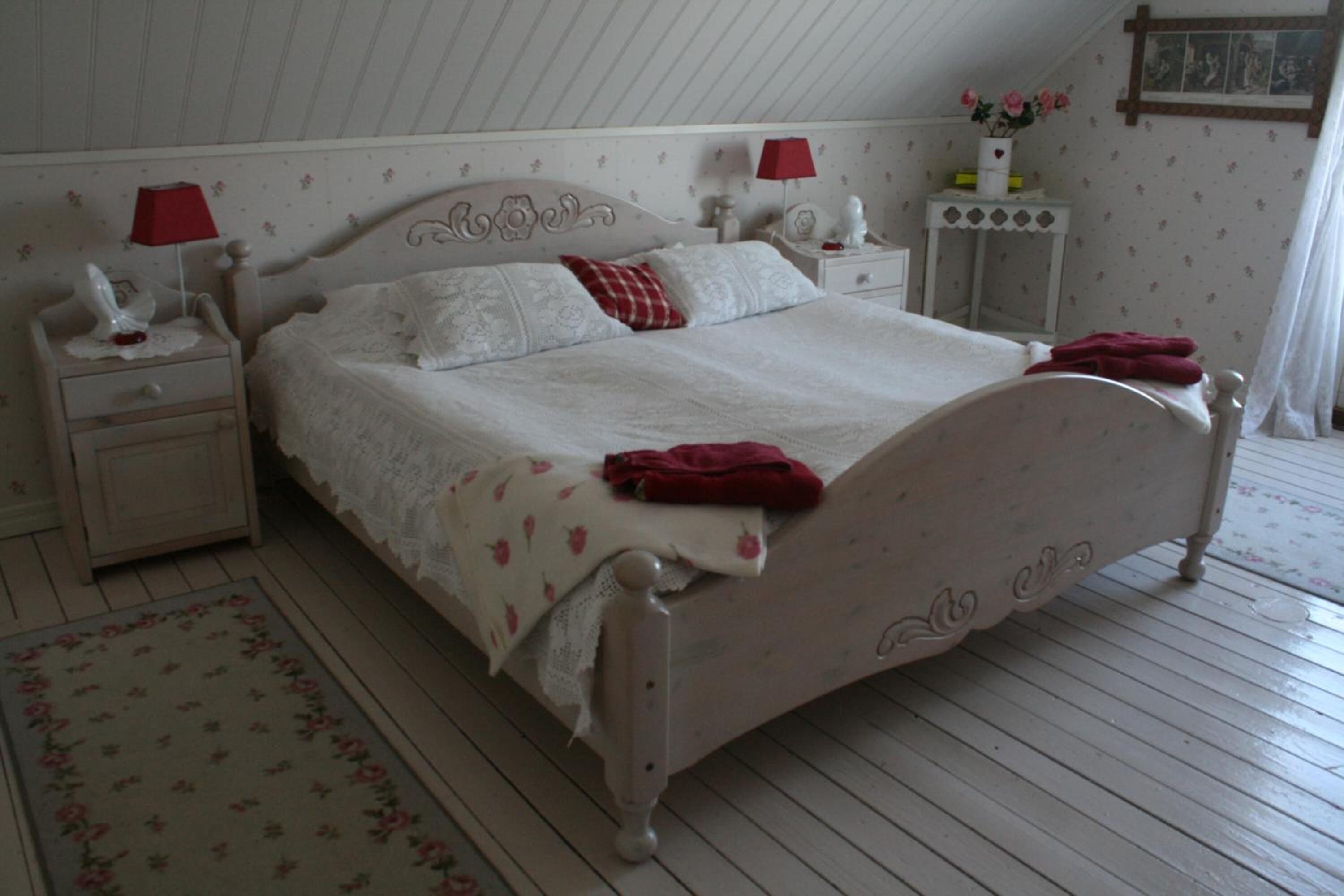 Un dormitorio con una cama con dosel blanco, una mesita de noche con lámpara, un aparador y una pequeña mesa con un jarrón de flores.