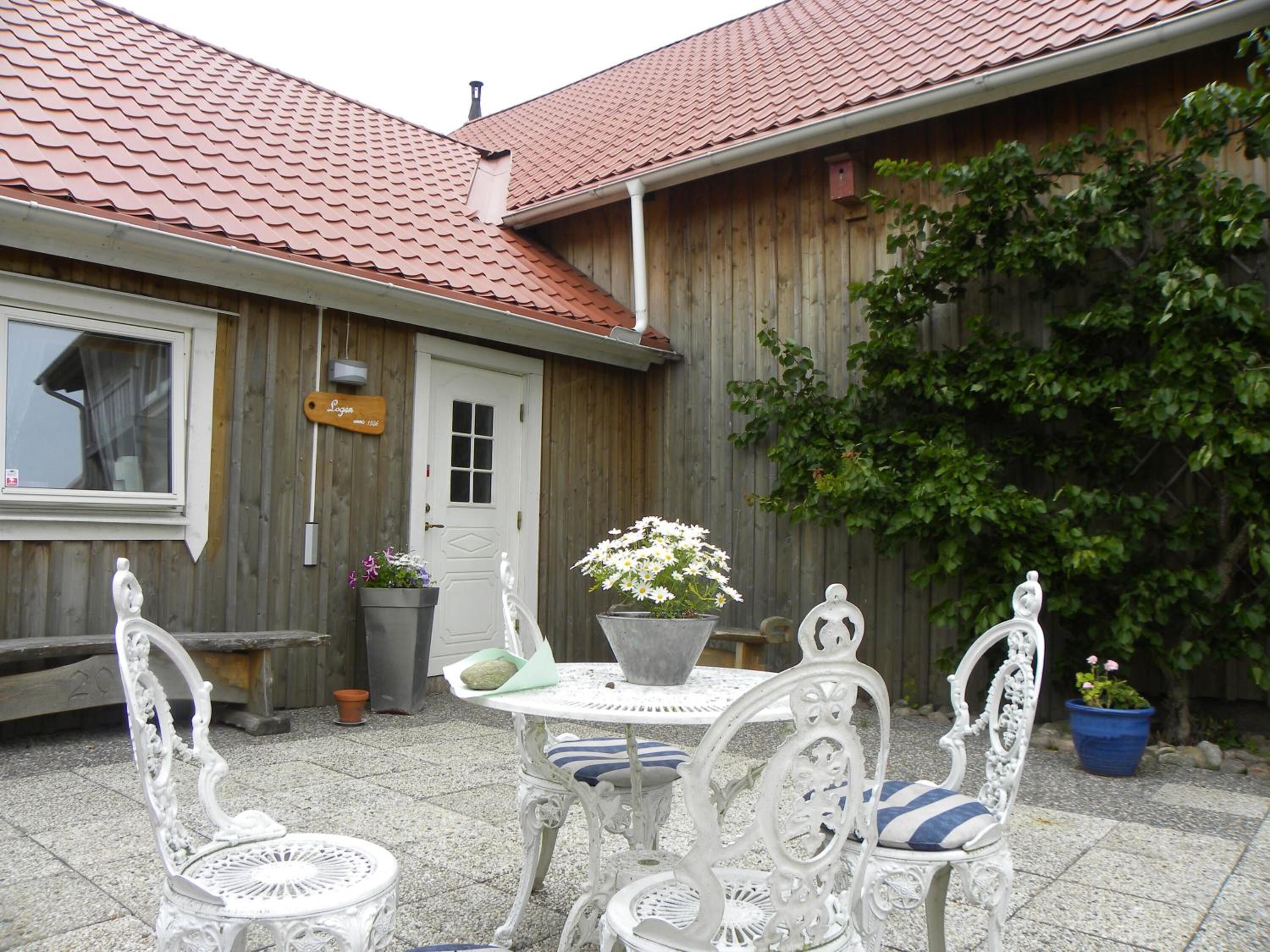 Ett trähus med ett rött tegeltak, en vit dörr och en liten trädgård med vita metallstolar och ett bord.