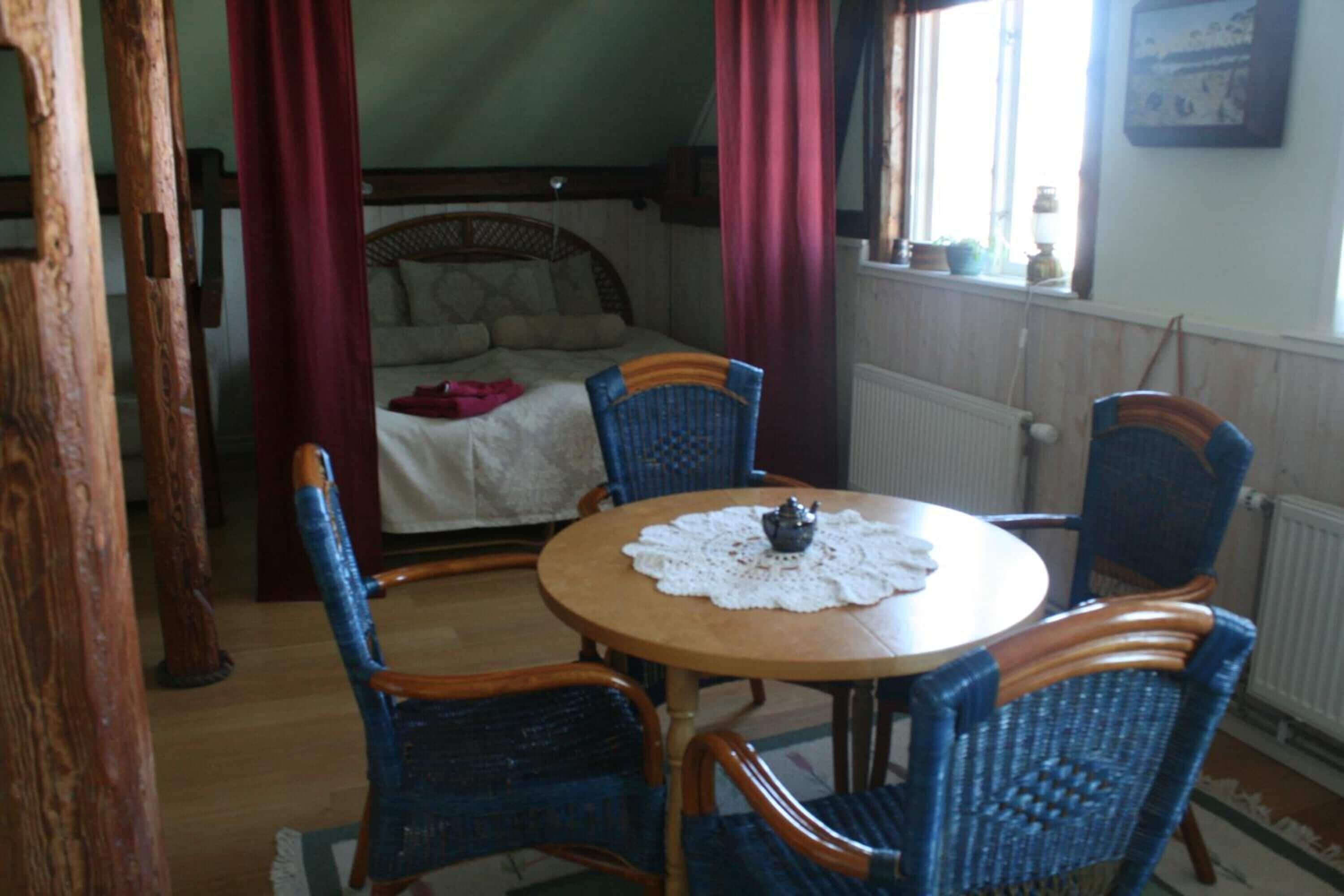 A room with a round wooden table, four blue wicker chairs, a bed with a white bedspread, and a window with curtains.