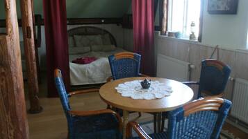 A room with a round wooden table, four blue wicker chairs, a bed with a white bedspread, and a window with curtains.