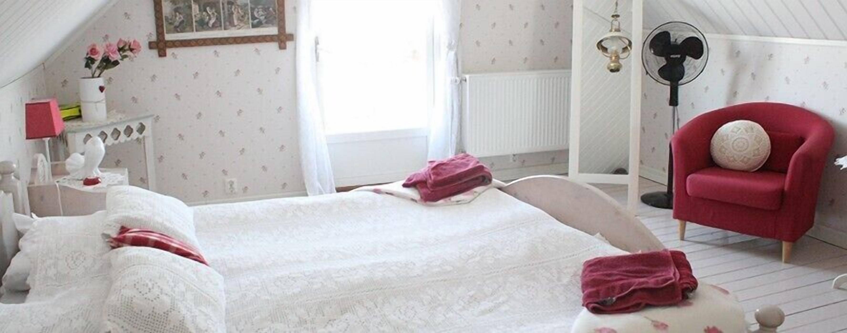 A bedroom with a large bed, a red armchair, a small table with a lamp, and a wall-mounted fan.